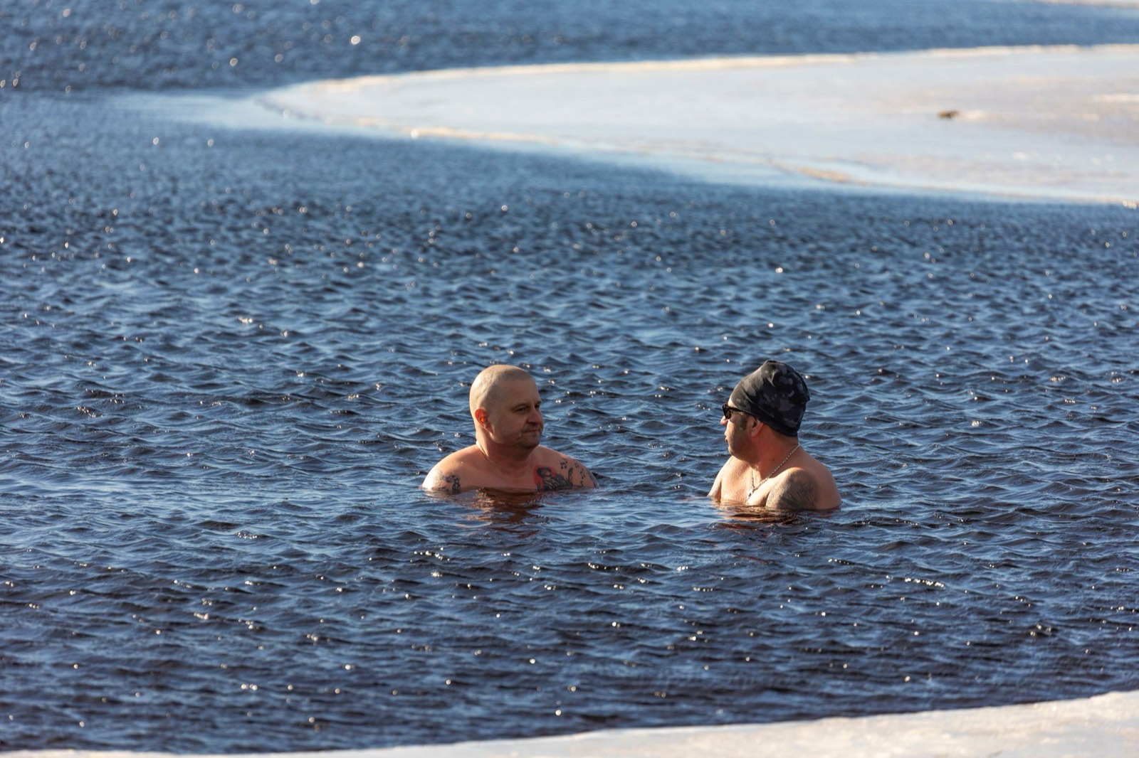 Two men stand shoulder-deep in dark, icy open water beside a shelf of ice, talking in winter sunlight.