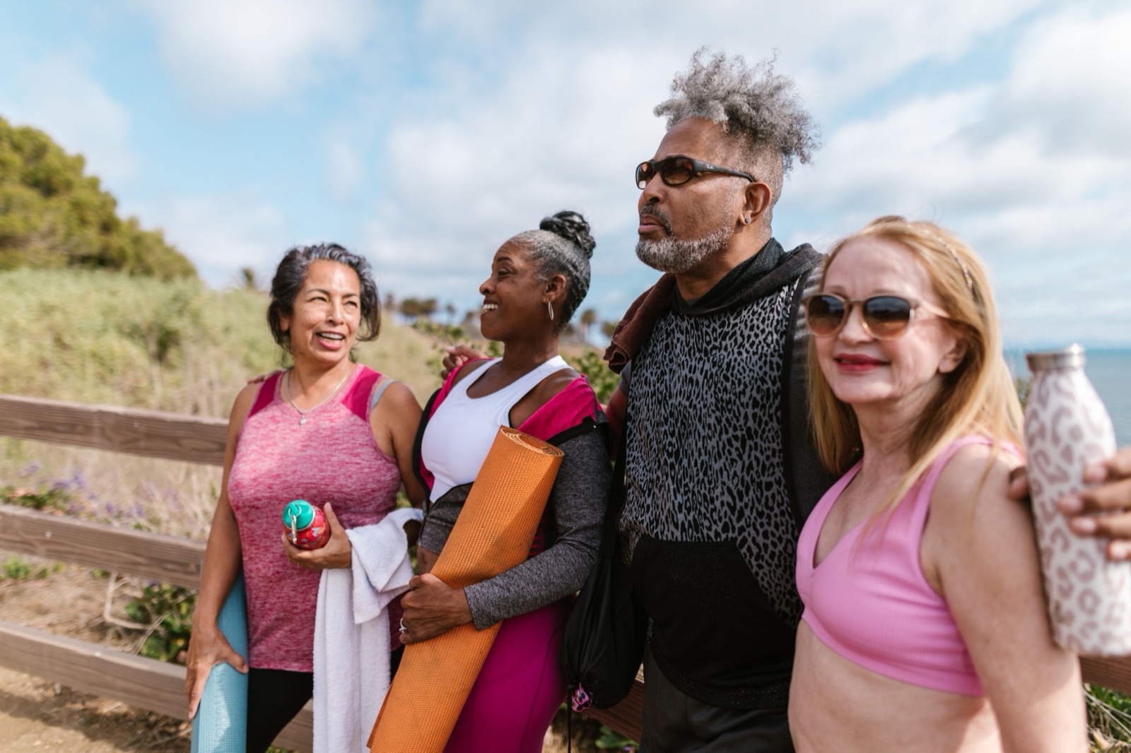 Four adults in workout clothes, carrying yoga mats and water bottles, stand together and chat outdoors on a sunny coastal path.