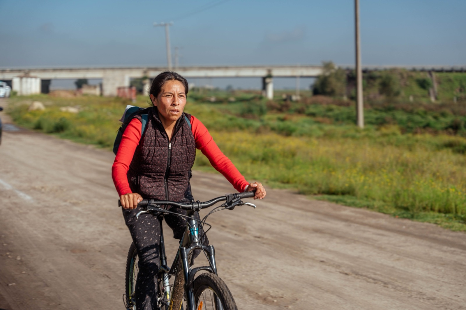 A woman in a red top and padded vest rides a bicycle along a dirt road in a grassy rural area.