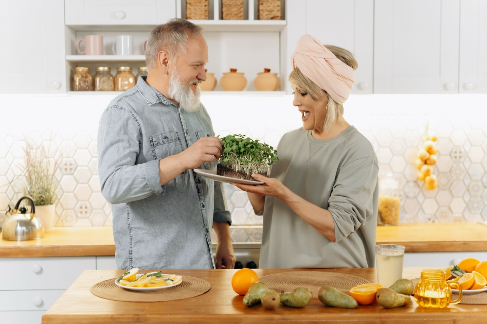 An older couple laugh together in a bright kitchen over a tray of fresh microgreens, with pears and oranges on the counter.
