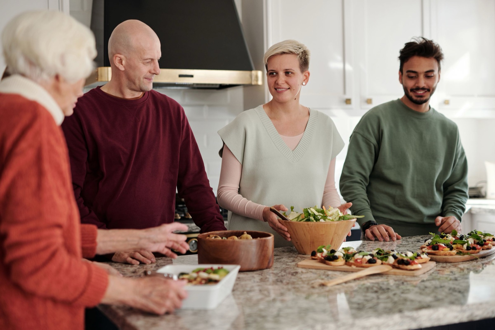 Four adults of different ages chat around a kitchen island as one tosses a big wooden bowl of salad beside trays of vegetable-topped toasts.