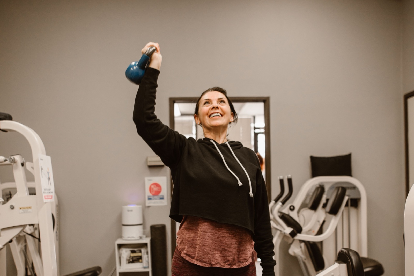 A smiling woman in a black hooded top lifts a blue kettlebell overhead with one arm in a small gym.