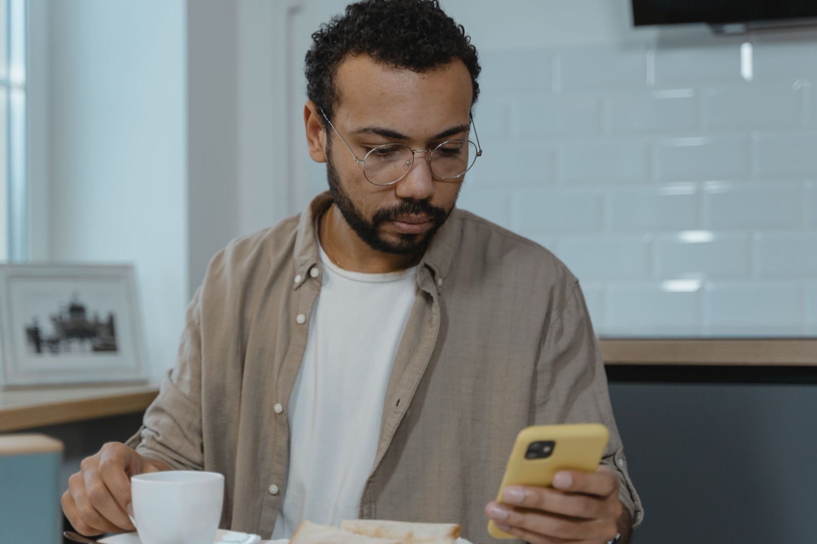 A bearded man in round glasses reads his phone at the kitchen table, a cup of coffee in his other hand.