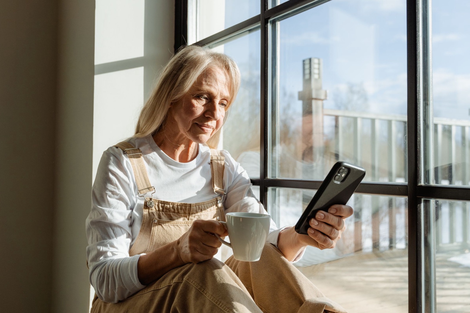 A woman with long fair hair sits by a large sunny window with a mug of coffee, reading on her phone.