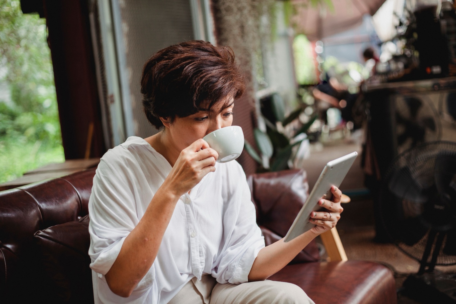 A woman with short hair sips coffee while reading on a tablet, seated on a leather couch.