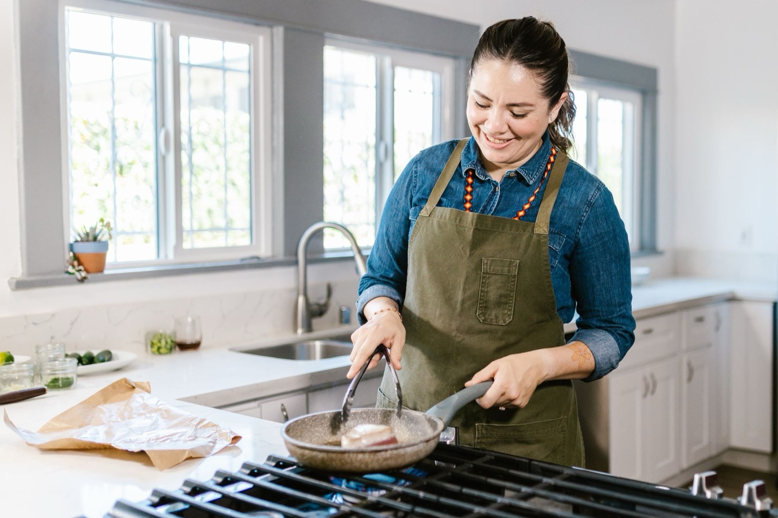 A smiling woman in a denim shirt and green apron turns food in a frying pan on a gas stovetop in a bright home kitchen.